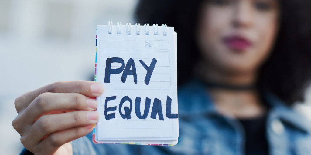 Stock image of a woman in the background, slightly blurred with her hand holding a note that says 'PAY EQUAL' in the foreground